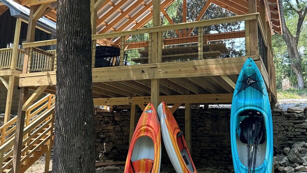Una cabaña de madera con balcón, una pared de piedra y kayaks apoyados.
