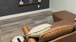 Una sala de estar moderna con un televisor de pared que muestra Paramount+, un sofá de cuero marrón con cojines, una pequeña mesa auxiliar de madera con un control remoto y objetos decorativos en estantes debajo del televisor.