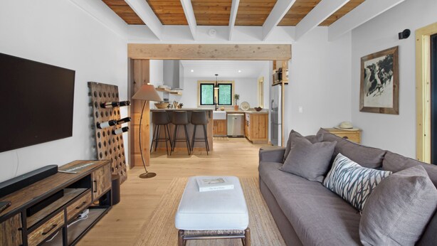A modern living room with a grey sofa, wooden ceiling beams, a kitchen in the background, and a TV on the left. - Ranch Retreat a mile from Warm Springs Base Lodge; New home just under an acre (Ketchum)