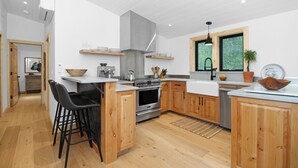 A modern kitchen with wooden cabinets, a central island, bar stools, and a large window. - Ranch Retreat a mile from Warm Springs Base Lodge; New home just under an acre (Ketchum)