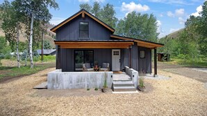A modern two-story house with a dark exterior, a wooden front porch, and a small outdoor seating area. - Ranch Retreat a mile from Warm Springs Base Lodge; New home just under an acre (Ketchum)
