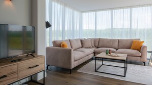 Un salon moderne avec un canapé d’angle, un téléviseur à écran plat, une table basse et une grande fenêtre ornée de rideaux légers.