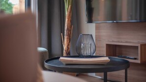 Ein modernes Wohnzimmer mit großem Flachbildfernseher, einem Holz-TV-Schrank, einem runden schwarzen Tisch und einer dekorativen Vase sowie einer großen Pflanze.