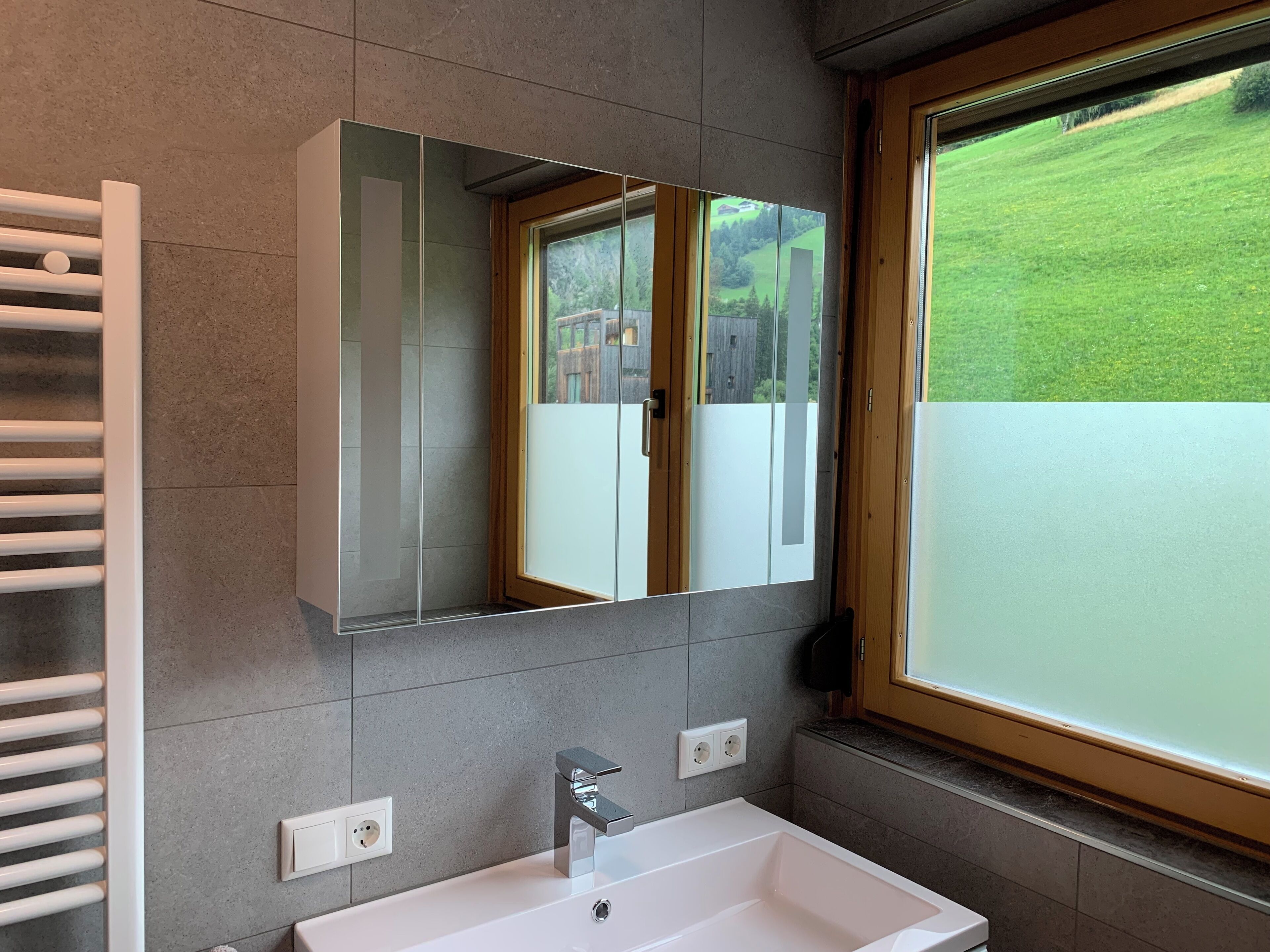 Ein modernes Badezimmer mit einem großen Spiegel, einem weißen Waschbecken und einem Fenster mit Blick auf eine grüne Landschaft.