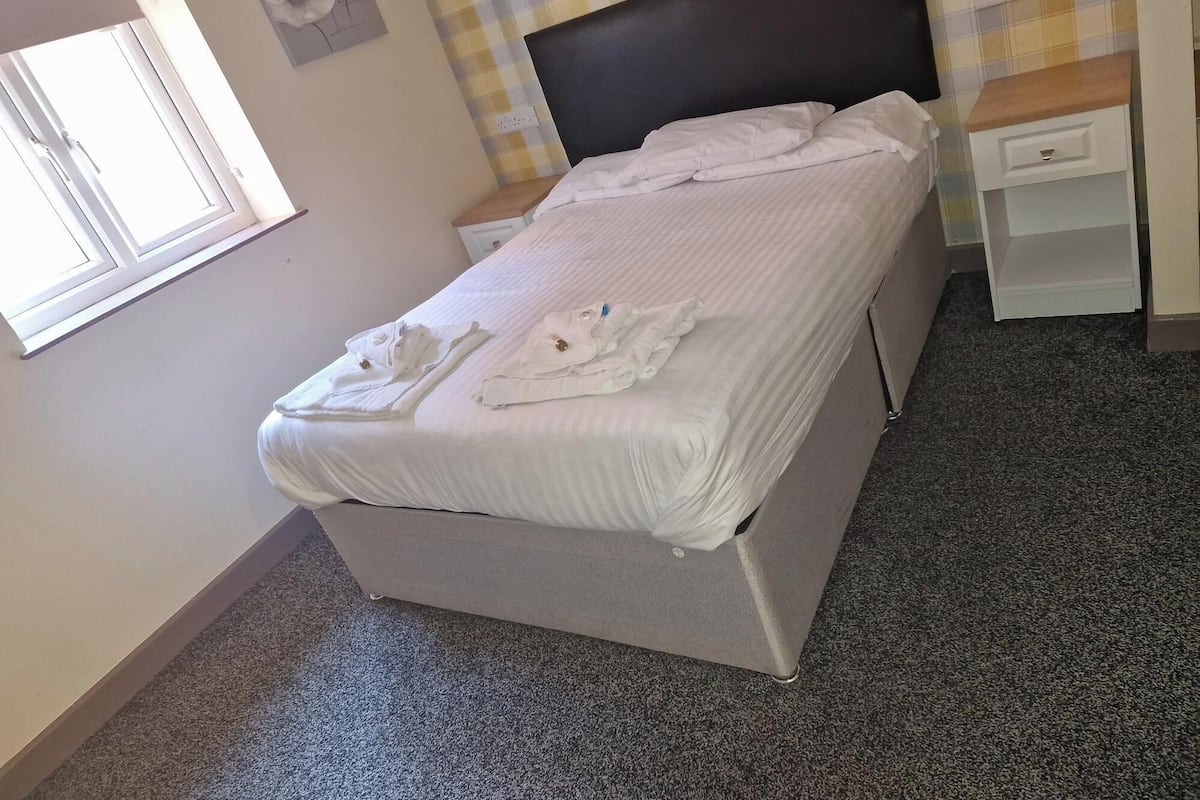 A neatly made bed with towels, a bedside table, a nightstand, and a wall with a floral painting.