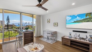 A spacious living room with a large window, a ceiling fan, a dining area, and a view of the beach.