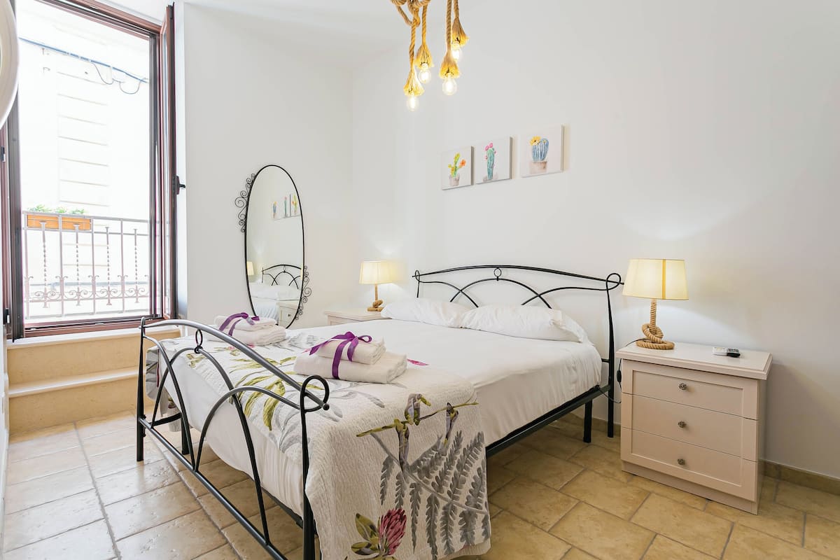 A bedroom with a bed, bedside tables, a mirror, and a window with a balcony.