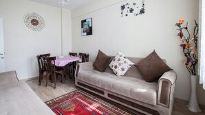 A living room with a beige sofa, two brown cushions, a patterned rug, a dining table with chairs, a clock, wall art, and a vase with flowers. - Convenient 3-Bedroom flat for 10, Near Blue Mosque, AC & WiFi (Istanbul)