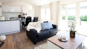 A modern kitchen with white cabinets, a dining area with a black sofa, and a wooden coffee table with a cake. - Nice Chalet Near the Golf Course in a Holiday Park (Velsen-Zuid)