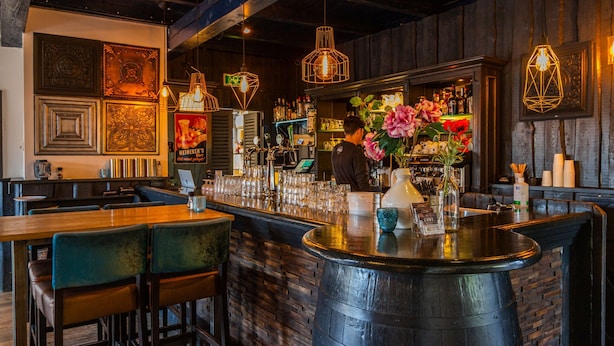 A bar area with a counter, stools, and hanging lights. There are decorative elements on the wall and a person working behind the bar. - Nice Chalet Near the Golf Course in a Holiday Park (Velsen-Zuid)