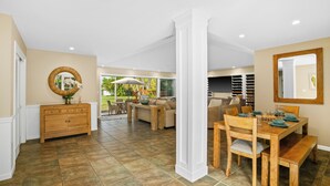 A spacious living area with a wooden dining table, a sofa, and a large window offering a view of the outdoors. - 3BR Poipu Kai home 5 min walk to beach and pool summer deals (KOLOA)
