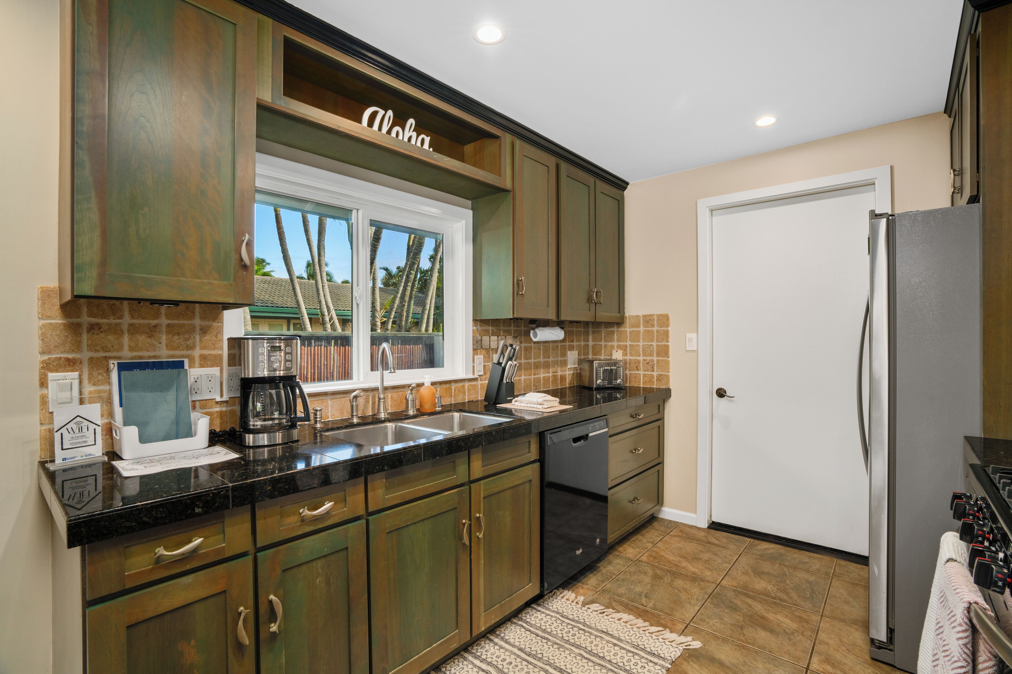 Una cocina con armarios de madera oscura, encimera negra, refrigerador de acero inoxidable y ventana con vista a palmeras.