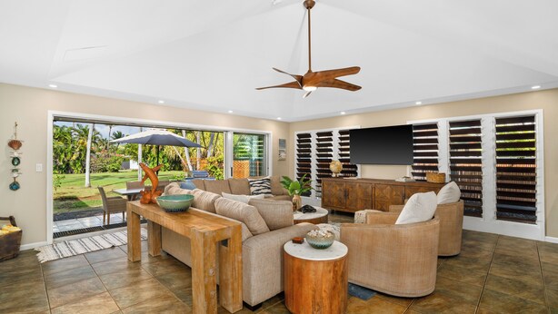 A spacious living room with a large sofa, a coffee table, and a dining area. There is a ceiling fan, a bookshelf, and a large window with a view of the outdoors. - 3BR Poipu Kai home 5 min walk to beach and pool summer deals (KOLOA)