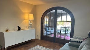 Een woonkamer met een bank, een bijzettafeltje, een lamp en uitzicht door een groot, rondbogig raam.