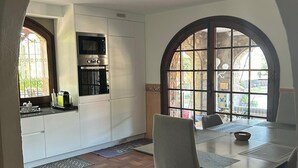 Een eethoek met een tafel en stoelen, een keuken met witte kasten en apparaten, en een uitzicht door een boogvormig raam.