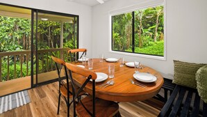 A dining area with a round table set for four, surrounded by chairs, with a view of lush greenery through large windows. - Summer Deal Princeville 3BR Kauai Townhome with AC and Parking (PRINCEVILLE)