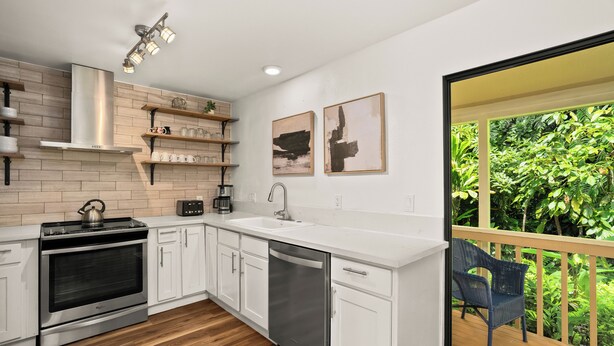 A modern kitchen with white cabinets, a stainless steel stove, and a dishwasher. There are two framed artworks on the wall and a chair by a sliding glass door leading to a balcony with greenery. - Summer Deal Princeville 3BR Kauai Townhome with AC and Parking (PRINCEVILLE)