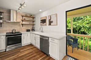 Een moderne keuken met witte kasten, een roestvrijstalen fornuis en een vaatwasser. Twee kunstwerken hangen aan de muur en er staat een stoel bij een schuifdeur die leidt naar een balkon met beplanting.