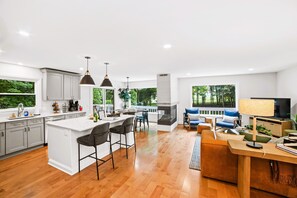Una cocina moderna con una isla central, taburetes de bar y un comedor con sofá y televisor.