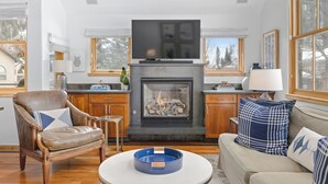 A living room with a fireplace, a television, wooden furniture, and a view of trees outside. - Owl Meadows-Curate Telluride-Fireplace, Views, Dog OK, Near Market, Lift 7 (Telluride)