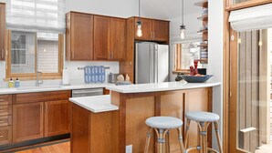 A modern kitchen with wooden cabinets, a stainless steel refrigerator, and a white countertop. There are two bar stools and a window with blinds. - Owl Meadows-Curate Telluride-Fireplace, Views, Dog OK, Near Market, Lift 7 (Telluride)