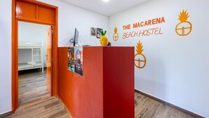 Un comptoir d’accueil avec un plan de travail rouge et une décoration représentant un ananas. Sur le mur, on peut lire une enseigne indiquant : « The Macarena Beach Hostel ».