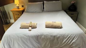 A neatly made bed with two towels and a bottle of lotion, a bedside lamp, and a vase with flowers on a nightstand.