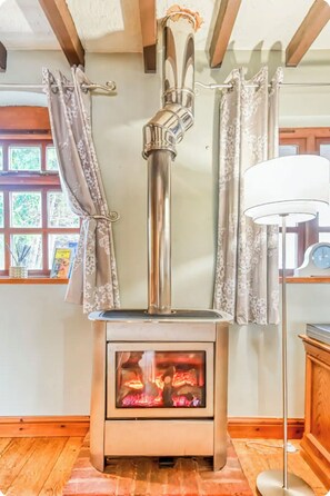 A wood-burning stove with a glass front, curtains, and a lamp in a room with wooden floors and a window. - The Hurst Stable, 1800 century Coach House offering luxurious amenities, Hottub  (Wheaton Aston)