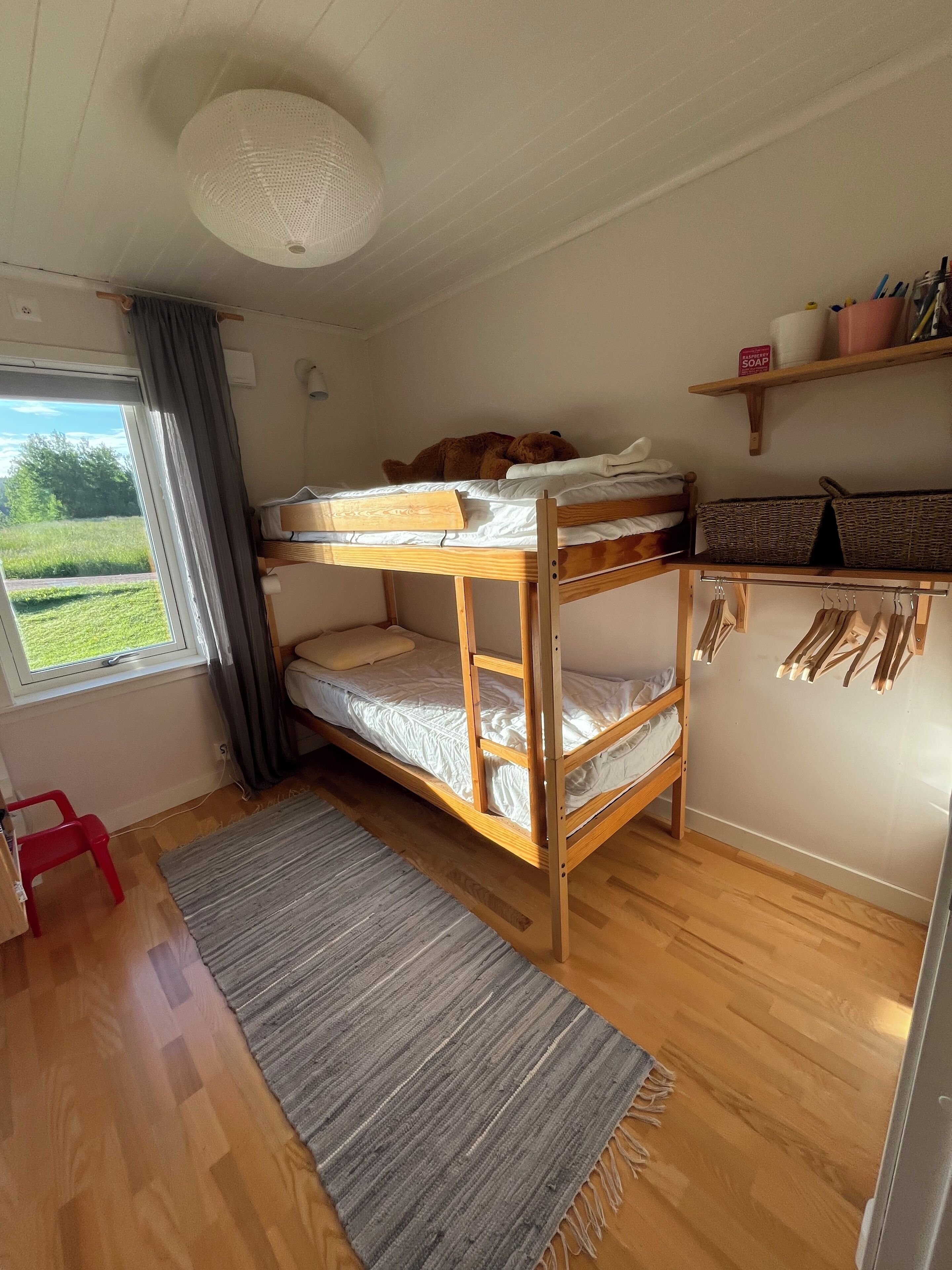 Une chambre avec un lit superposé, une fenêtre, une chaise rouge, un tapis gris et une étagère avec des paniers.
