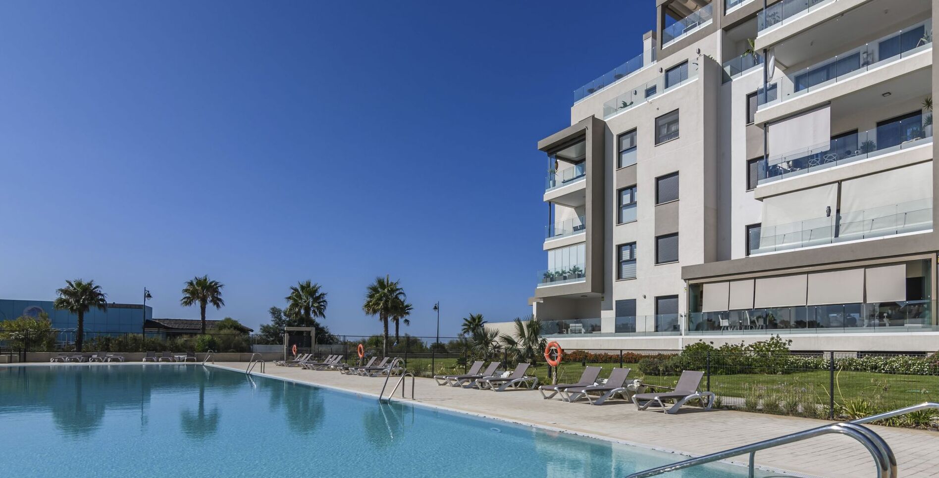A modern hotel with a swimming pool, sun loungers, and a clear blue sky.