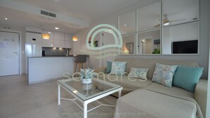 A modern living room with a glass coffee table, a sofa with patterned cushions, and a kitchen area with white cabinets and a built-in oven. - Los Flamencos 5004 - Two Bedroom Apartment, Sleeps 5 (Punta del moral)