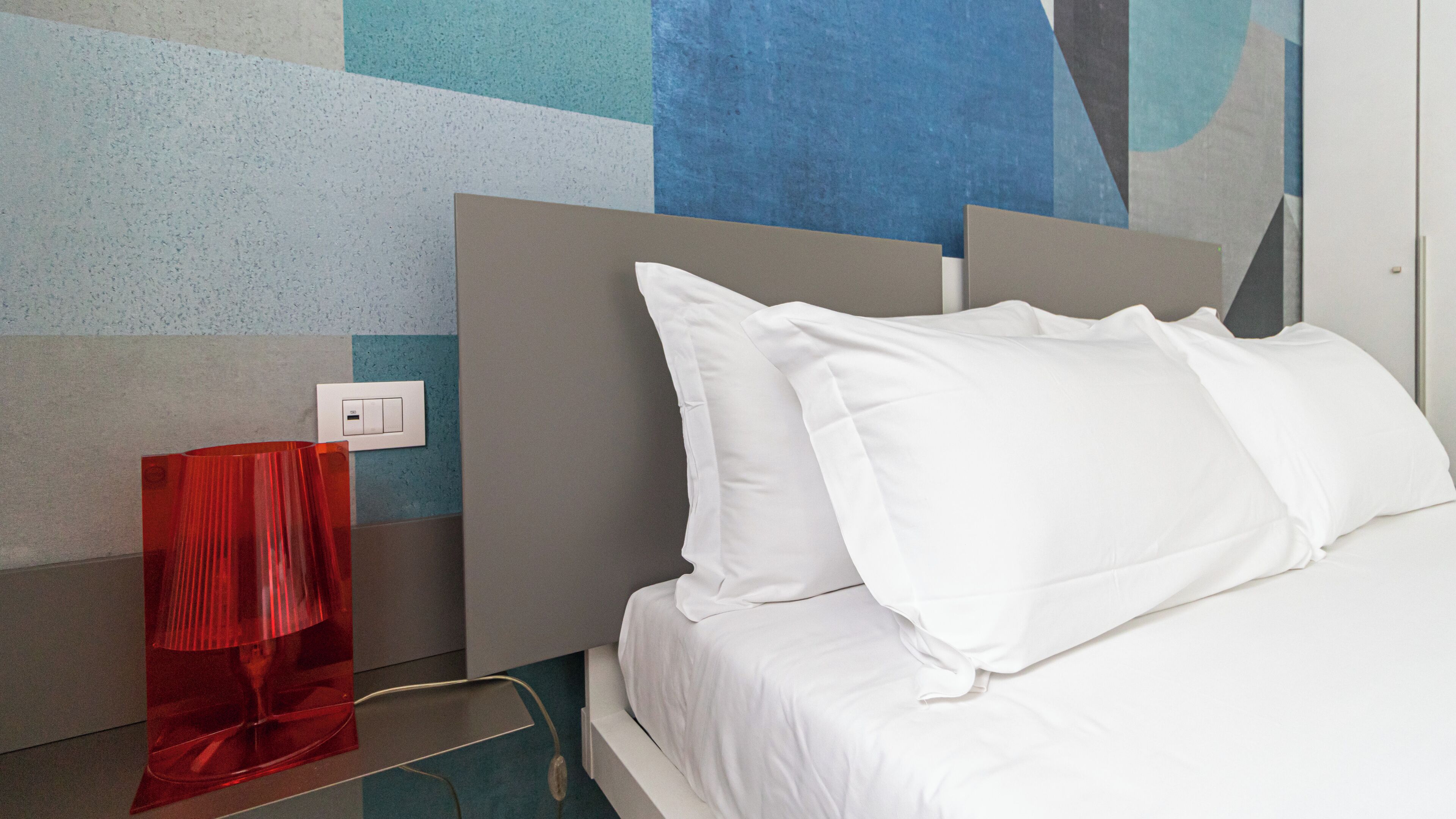 A hotel room with a bed, white bedding, a red lamp, and a wall with a geometric pattern.