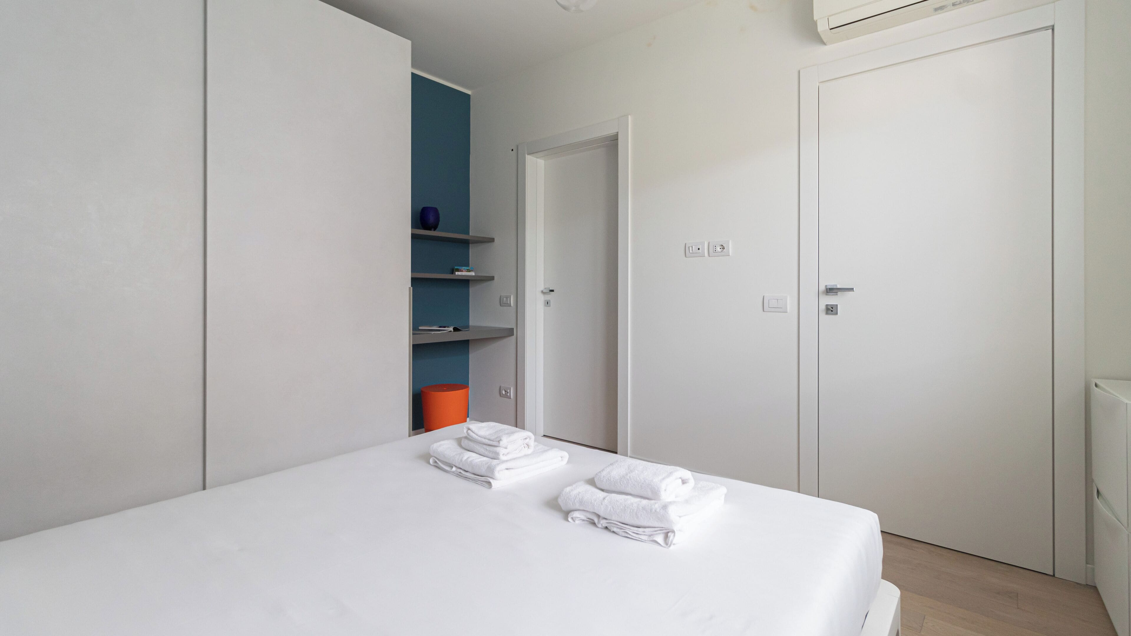 Een moderne slaapkamer met een bed, witte muren, een blauwe wandplank en een witte deur.