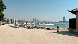A beach with rows of sun loungers and umbrellas, a clear view of the city skyline in the background, and a calm sea.