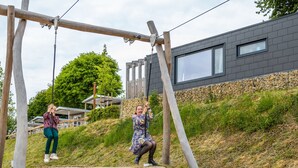 A wooden swing set in a park with a modern building in the background. - Comfy Chalet With Cooking Island in Limburg (Gulpen)