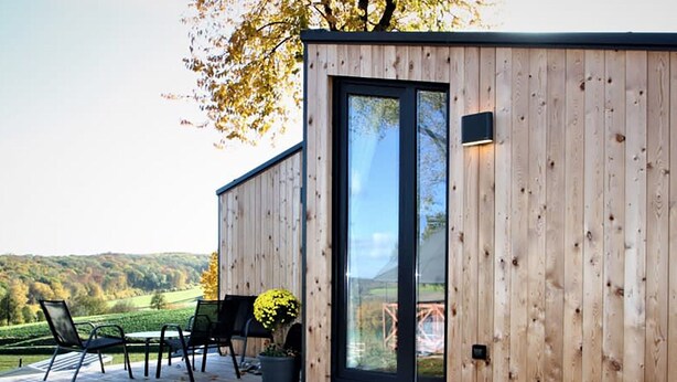 A wooden cabin with a glass door, a deck with chairs, and a view of the countryside. - Comfy Chalet With Cooking Island in Limburg (Gulpen)
