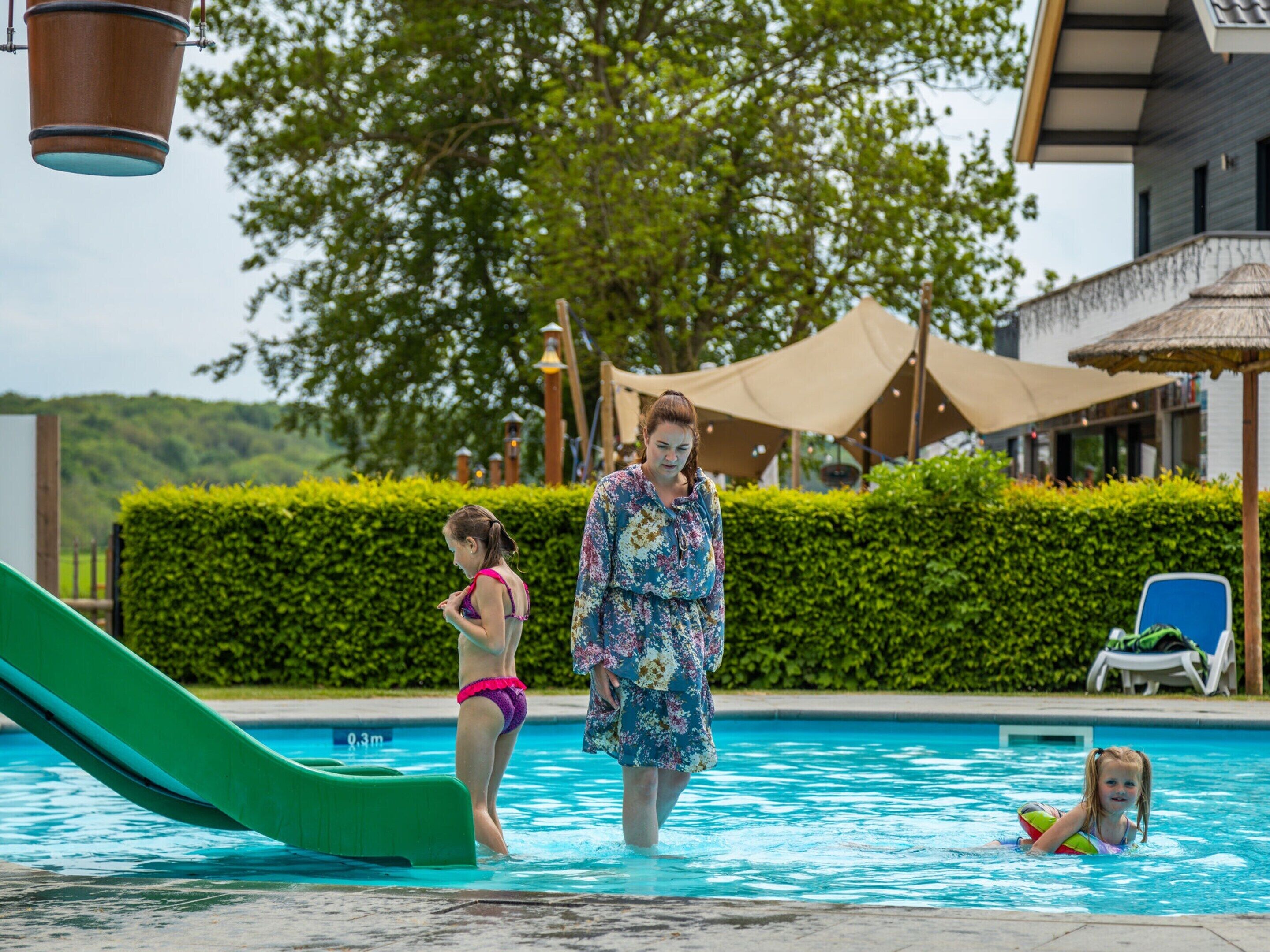 Une piscine avec un toboggan, entourée de verdure et bordée d’un bâtiment à toiture de chaume.