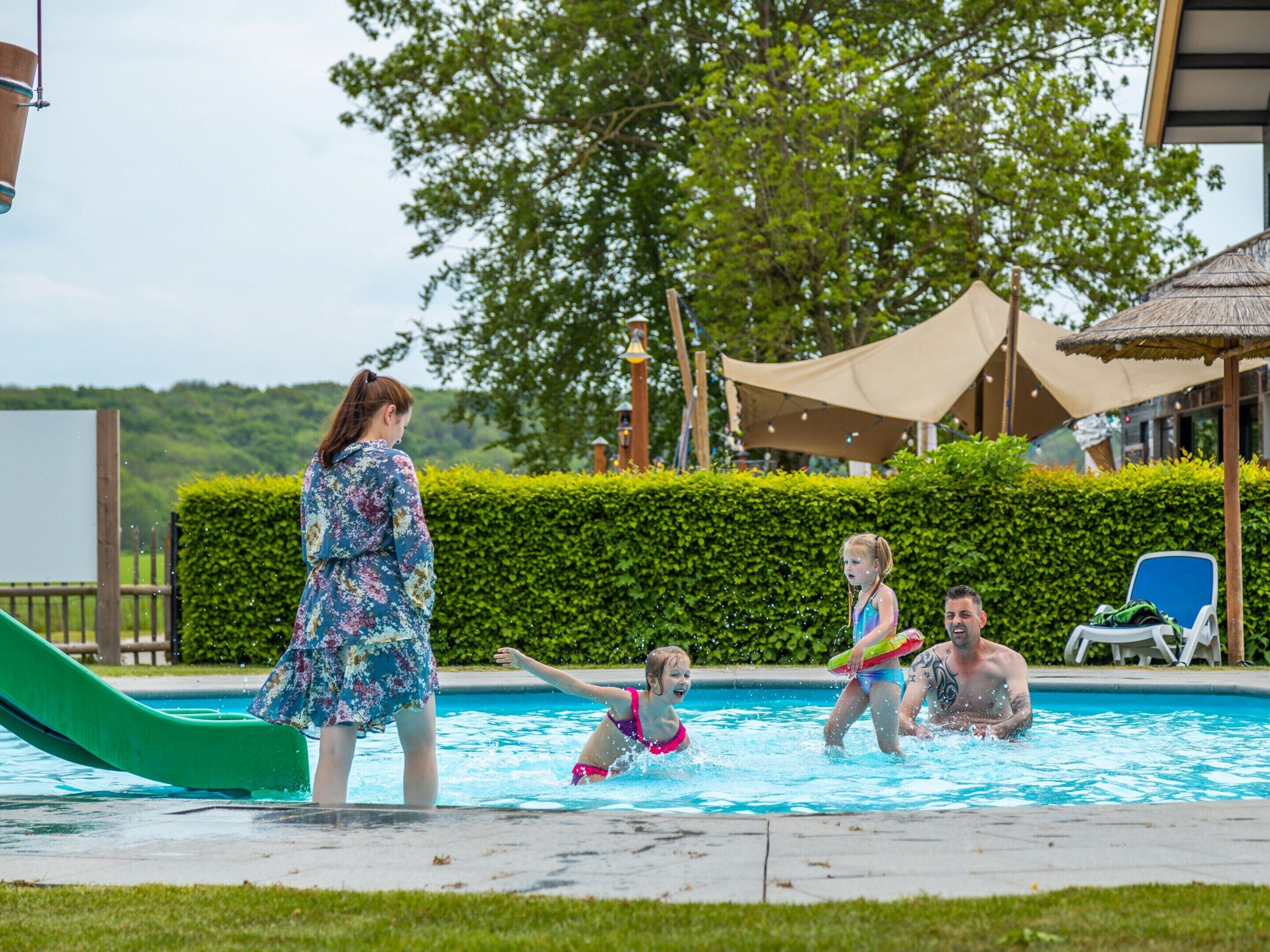 A swimming pool with a slide, surrounded by greenery and a building with a thatched roof.