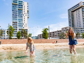 A person in a blue dress and a striped shirt playing in shallow water with splashes around them, with tall buildings in the background. - Well-kept Chalet With Dishwasher (Nieuwerkerk aan den IJssel)