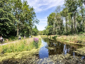 A calm river with dense trees on both sides, a narrow path on the left, and a few people enjoying the outdoors. - Well-kept Chalet With Dishwasher (Nieuwerkerk aan den IJssel)