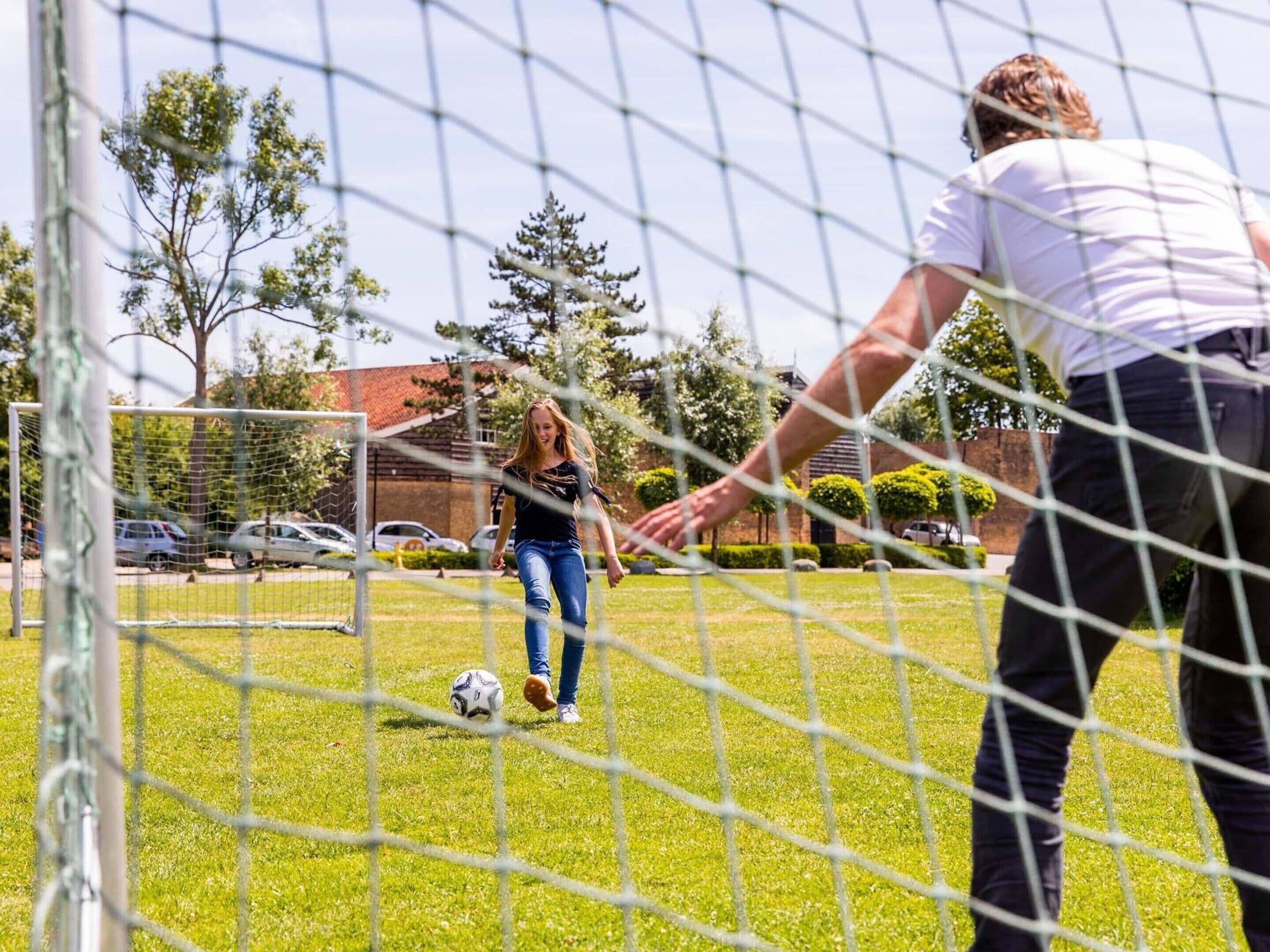 Ein Fußballtor mit Netz, ein Fußball auf dem Rasen und eine Person im Hintergrund.
