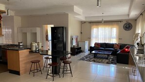 A modern kitchen with a central island, a dining area with stools, a living room with a sofa and a coffee table, and a kitchen counter with a refrigerator and a TV.