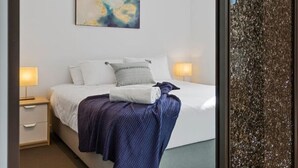 Una cama bien hecha con una colcha azul, almohadas blancas y un cuadro encima. Hay dos mesitas de noche con lámparas.