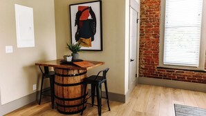 Une pièce avec une table en bois en forme de tonneau, deux chaises noires, un tableau abstrait encadré, une plante en pot et une fenêtre avec des stores.