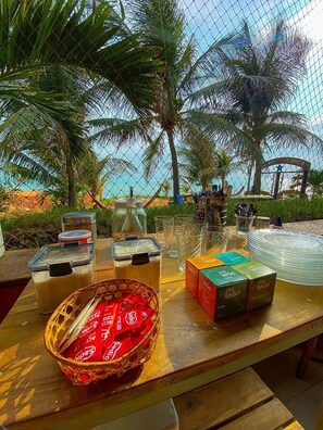 A table set up with various food items and drinks, surrounded by palm trees and a view of the ocean. - Raio de Sol Residence (Aracati)