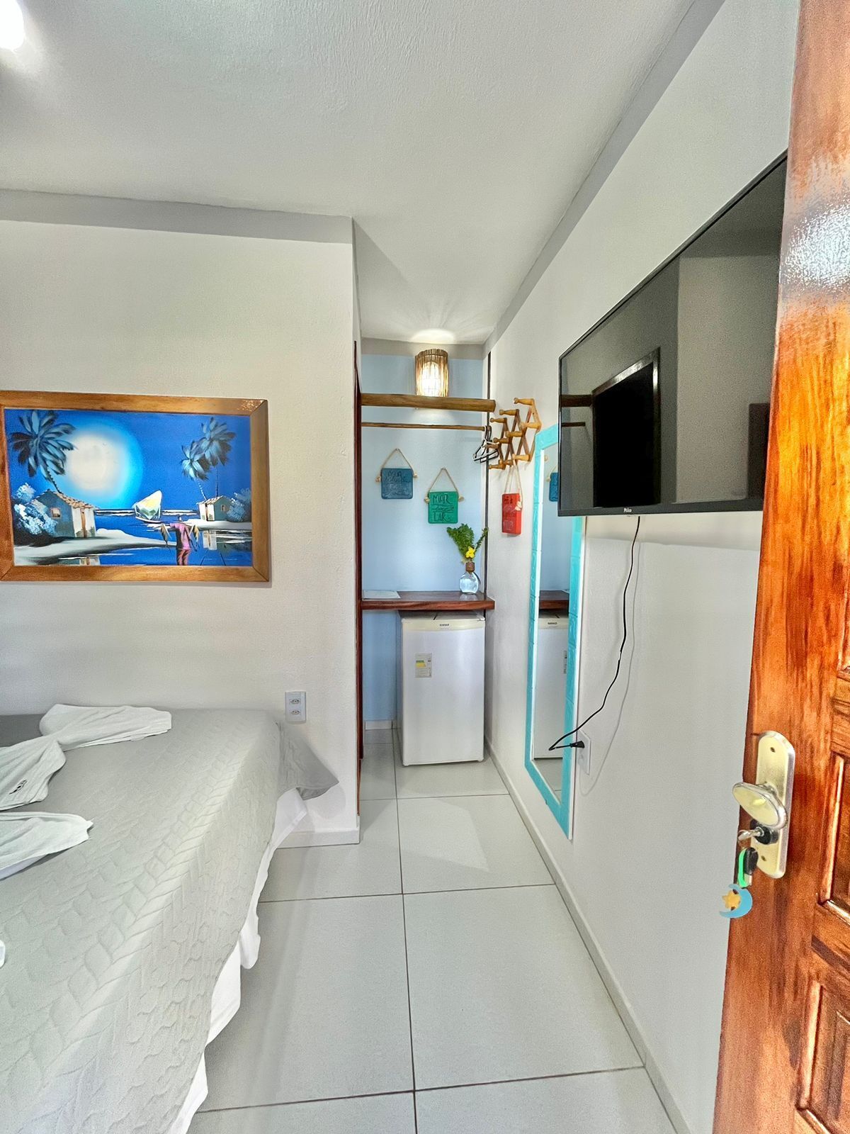 Quarto de hotel compacto com cama, pequeno espaço de cozinha, TV fixada na parede e um quadro na parede.