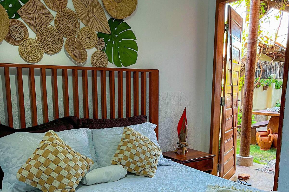 A neatly made bed with a checkered pillow, a woven wall hanging, and a towel on the floor.