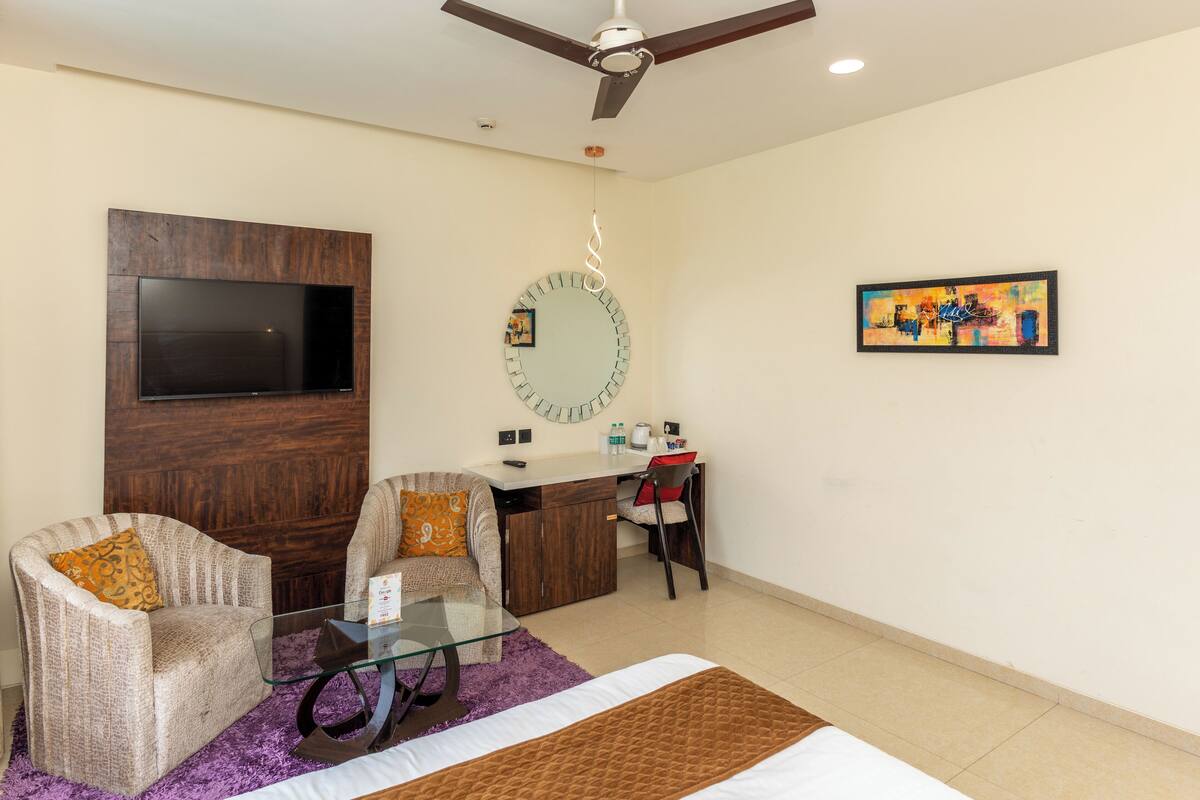 A modern hotel room with a bed, a glass coffee table, two armchairs, a desk with a chair, a TV mounted on the wall, a round mirror, and a painting.