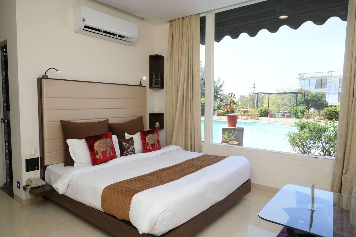 A hotel room with a large bed, a small table, a phone, and a view of a swimming pool and greenery.
