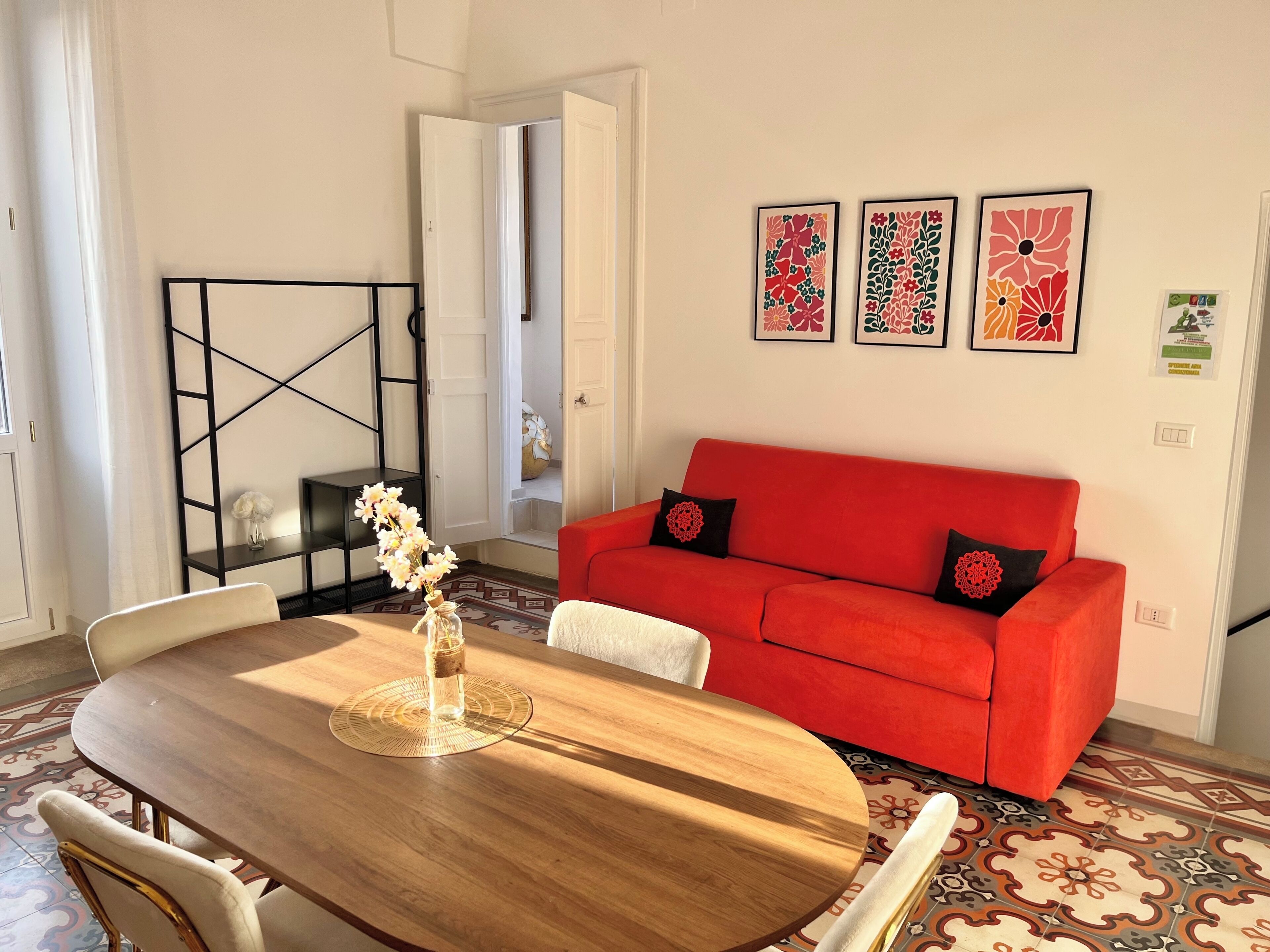 Un salon avec une table à manger en bois, un canapé rouge et trois œuvres d’art florales encadrées accrochées au mur.